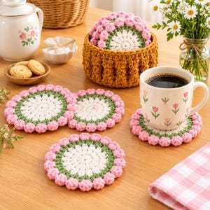 May include: A set of round, crocheted coasters featuring a white center, a green middle ring, and a pink scalloped edge. The coasters are displayed alongside a matching mustard-colored crocheted basket, a ceramic mug with a tulip pattern, and cookies.