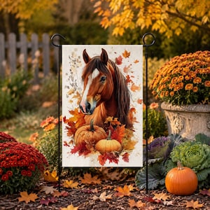 May include: A rectangular garden flag featuring a watercolor illustration of a brown horse head surrounded by vibrant orange and red autumn leaves and two pumpkins. The flag is suspended on a black metal stand in an outdoor garden setting.