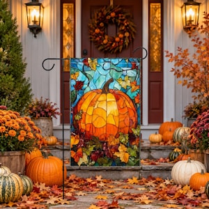 May include: A rectangular garden flag featuring a stained glass style illustration of a large orange pumpkin surrounded by colorful autumn leaves against a blue background, displayed on a black metal stand in front of a house entrance.