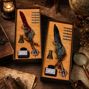 Op de afbeelding: Twee rechthoekige geschenkdozen met ganzenveren versierd met metalen tandwielen en vleugels. Eén veer is donkerrood, de andere zwart met gouden spikkels. Elke set bevat een flesje zwarte inkt, een metalen penhouder en vijf verwisselbare penpunten.