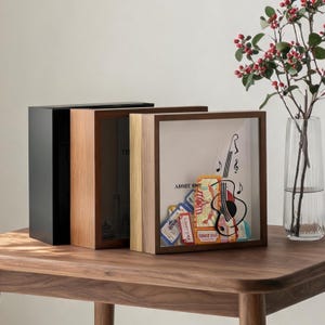 May include: Three square shadow box frames in black, light wood, and dark wood finishes. The front-most frame contains a collection of colourful ticket stubs and a black line drawing of a guitar with musical notes on the glass front.