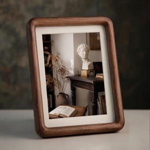 May include: A rectangular dark brown wooden picture frame with rounded corners and a cream-colored mat. The frame features the name Elena engraved in cursive script on the bottom edge. It displays a photograph of a bust, a book, and a vase.