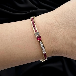 May include: A gold hinged bangle bracelet featuring a row of square-cut red rubies, a central pear-shaped ruby, and a series of round brilliant-cut diamonds. The design includes a small section of baguette-cut diamonds near the centre stone.