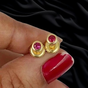 May include: A pair of gold stud earrings featuring round, vibrant pinkish-red gemstones set in a polished gold bezel. The earrings have a screw-back mechanism with a textured, circular base, showcasing a bright metallic finish and clear gemstone facets.
