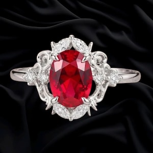 May include: A silver ring featuring a large oval-cut red gemstone at the center. The setting is adorned with smaller marquise and round-cut clear stones arranged in an intricate, decorative metal pattern surrounding the central red stone.