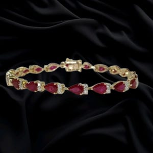May include: A gold bracelet featuring a series of pear-shaped red rubies alternating with small clear stones. The links are connected in a continuous circular design with a secure clasp mechanism visible at the top of the jewelry piece.