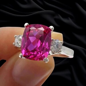 Puede incluir: Un anillo de plata con una gran piedra preciosa de color rosa vibrante y corte cojín como pieza central, flanqueada por dos piedras blancas redondas y transparentes a cada lado. La banda es de plata pulida, creando un contraste metálico.