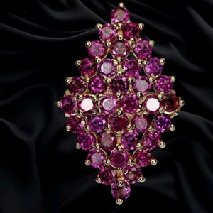 Puede incluir: Un anillo de oro con un gran grupo en forma de diamante compuesto por numerosas piedras preciosas redondas, facetadas y de color rojo rosado intenso. Las piedras están engastadas estrechamente en un marco de metal, creando un patrón denso y brillante.
