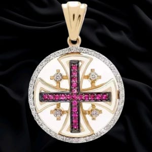 May include: A circular gold pendant featuring a Jerusalem cross design. The central cross is inlaid with vibrant pink stones, while four smaller white stones are positioned in the quadrants. The outer edge is bordered by small white stones.