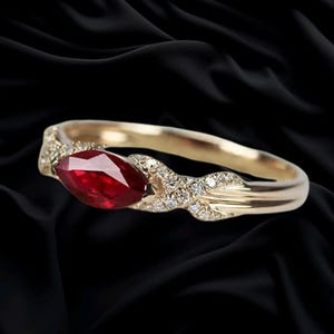 May include: A gold ring featuring a central marquise-cut red ruby gemstone. The band is accented with small, round, clear pavé-set diamonds along the sides, leading into a smooth, polished gold shank that tapers toward the back of the ring.