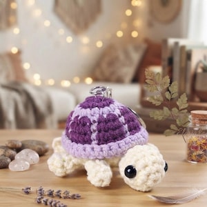May include: A crochet turtle keyring featuring a cream-coloured body and head with large black eyes. The turtle shell is crafted with a dark purple centre and a light lavender border. A silver metal keyring is attached to the top of the shell.