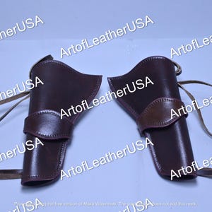 Op de afbeelding: Een paar donkerbruine lederen holsters met een gebogen bovenrand, een horizontale band in het midden en bevestigde lederen koorden. De tekst ArtofLeatherUSA is herhaald over de afbeelding. Deze holsters zijn bedoeld voor het dragen van voorwerpen.