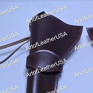 Pode incluir: Um coldre de couro castanho escuro com acabamento liso, costuras visíveis ao longo das bordas e uma tira larga no centro. Um cordão de couro fino estende-se da lateral do coldre, que está marcado com o texto ArtofLeatherUSA.