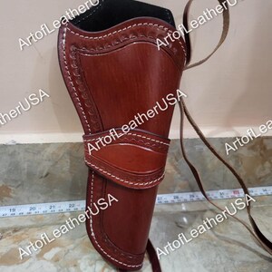 May include: A brown leather holster featuring decorative stamped edges, white stitching, and a black interior lining. A thin leather strap is attached to the side. A white measuring tape is visible, showing a length of approximately 12 inches.