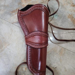 Pode incluir: Um coldre de couro castanho com padrões decorativos em relevo nas bordas, costuras brancas e tiras longas de couro. O coldre tem uma forma estruturada e curva com uma faixa horizontal central e um acabamento exterior liso e polido.