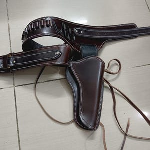 May include: A dark brown leather gun holster and belt set featuring a metallic buckle, white contrast stitching, and a series of small loops for ammunition. The belt displays the embossed letters WBH and includes long leather tie straps.