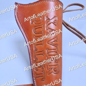 May include: A tan leather holster featuring white stitching along the edges and the embossed name XAVIOR QUILLET vertically on the front surface. The holster includes attached leather straps extending from the sides for secure fastening.