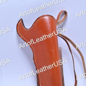 May include: A tan leather holster featuring white contrast stitching along the edges and a long, thin leather strap attached to the side. The holster has a curved top opening and is displayed on a small wooden stand with the text ArtofLeatherUSA.