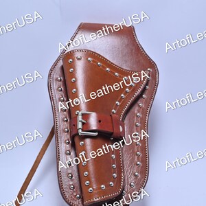 Pode incluir: Um coldre de couro castanho com uma borda decorativa de tachas metálicas prateadas. O coldre inclui uma correia central de couro com uma fivela metálica prateada para prender um objeto. Um cordão de couro longo e fino estende-se pelo lado.