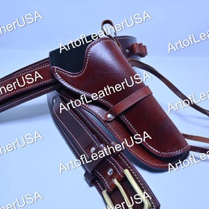 May include: A dark brown leather gun holster attached to a matching leather belt with a brass-toned metal buckle. The holster features white contrast stitching, a secure strap across the front, and a series of loops for ammunition storage.