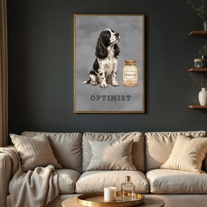 Op de afbeelding: Een ingelijste wandkunstprint met een zwart-witte spaniël die naast een glazen pot met het label TREATS zit. Het woord OPTIMIST staat in dikke grijze letters onder de hond gedrukt. De achtergrond is grijs met een subtiele wolkachtige textuur.
