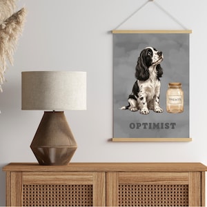 May include: A vertical wall hanging poster featuring a black and white spaniel dog sitting next to a glass jar labelled TREATS. Below the dog, the word OPTIMIST is printed in bold grey letters. The poster is held by light wood rails at the top and bottom.