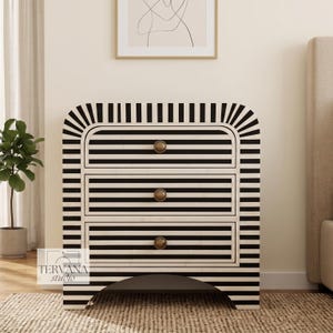May include: A three-drawer chest featuring a black and white striped pattern across the entire exterior. The piece has a rounded top edge, a curved base cutout, and three circular brass-toned knobs centered on each drawer front.