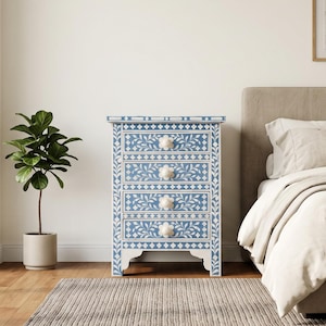 May include: A four-drawer chest featuring a blue and white floral inlay pattern. The rectangular piece has a flat top, straight sides, and four white flower-shaped knobs. The frame is decorated with geometric borders and leaf motifs.
