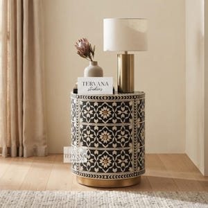 Puede incluir: Una mesita de noche cilíndrica con un patrón floral geométrico incrustado en blanco y negro, tres cajones con pequeños pomos dorados y una base dorada. Una lámpara dorada con pantalla blanca y un jarrón con una flor están encima. Texto TERVANA studios.