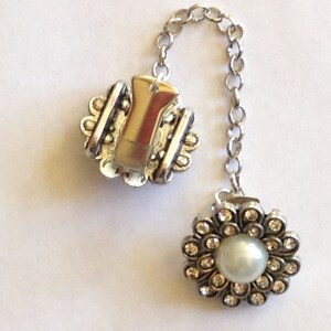 May include: A silver-toned brooch with a chain, featuring a floral design with a central pearl and surrounding clear crystals. The other end has a clip with a decorative edge.