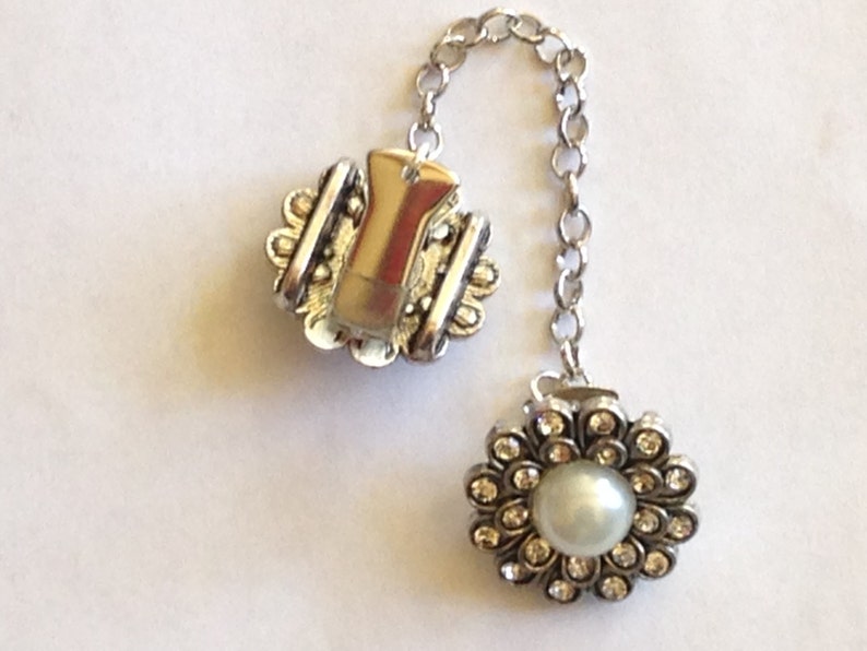 May include: A silver-toned brooch with a chain, featuring a floral design with a central pearl and surrounding clear crystals. The other end has a clip with a decorative edge.