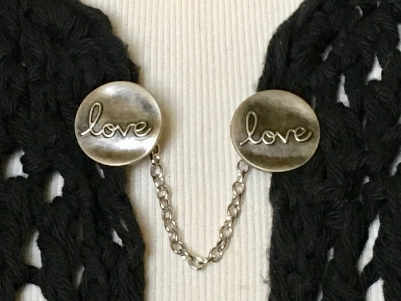 May include: A pair of silver tone scarf clips with a chain connecting them. Each clip features a round, slightly domed design with the word "love" embossed in a cursive font.