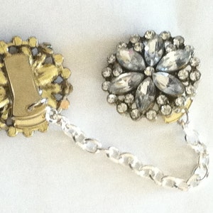 May include: A pair of vintage rhinestone and gold tone dress clips with a silver chain connecting them. The clips are round and feature a floral design with clear rhinestones.