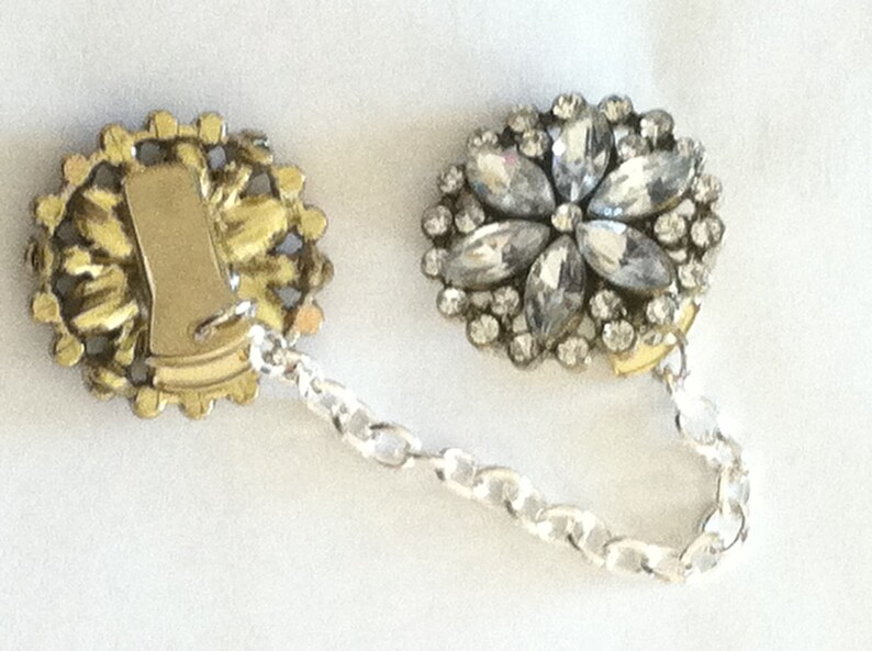 May include: A pair of vintage rhinestone and gold tone dress clips with a silver chain connecting them. The clips are round and feature a floral design with clear rhinestones.