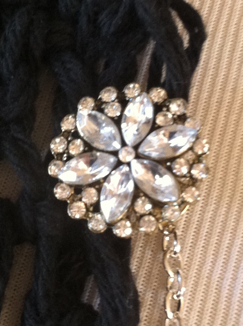 May include: A silver flower-shaped brooch with clear rhinestones. The brooch is attached to a silver chain.