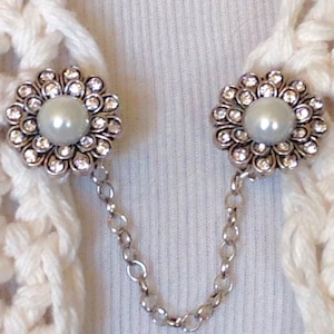 May include: A pair of silver-toned sweater clips with a floral design. Each clip features a central pearl surrounded by clear crystals. The clips are connected by a delicate silver chain, ideal for securing a cardigan or shawl.