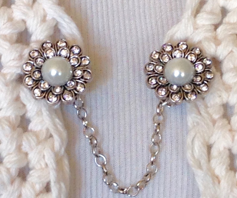 May include: A pair of silver-toned sweater clips with a floral design. Each clip features a central pearl surrounded by clear crystals. The clips are connected by a delicate silver chain, ideal for securing a cardigan or shawl.