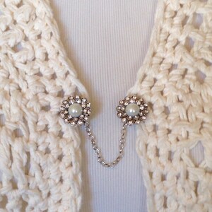 May include: Cream-colored crochet shawl with two decorative silver-toned flower-shaped clasps. Each clasp features a central pearl surrounded by clear rhinestones. A delicate silver chain connects the clasps.