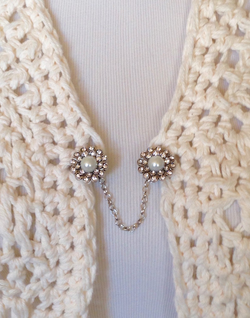 May include: Cream-colored crochet shawl with two decorative silver-toned flower-shaped clasps. Each clasp features a central pearl surrounded by clear rhinestones. A delicate silver chain connects the clasps.