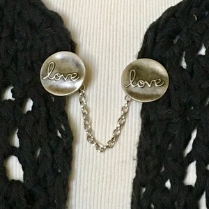 May include: A pair of silver tone metal clasps with a chain connecting them. Each clasp has the word "love" engraved on it.