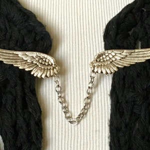 May include: A silver-coloured wing-shaped brooch with detailed feathering, linked by a fine chain. The brooch is set against black fabric and a white ribbed surface, creating a striking contrast.