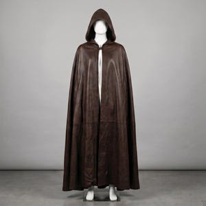 May include: A floor-length dark brown leather cloak featuring a deep hood and a single metal clasp closure at the neck. The garment drapes in long, vertical folds from the shoulders to the floor, covering the torso and legs completely.