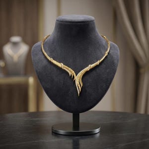 Puede incluir: Un collar de oro con una pieza central en forma de V con superficies metálicas texturizadas y piedras claras incrustadas. El diseño consta de dos ramas curvas simétricas que se encuentran en el centro, creando una forma puntiaguda.