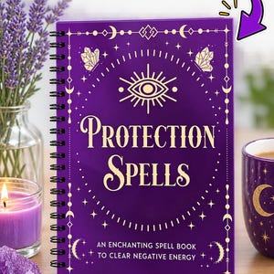 May include: A purple spiral-bound paperback book titled Protection Spells: An Enchanting Spell Book to Clear Negative Energy. The cover features gold celestial and eye motifs, stars, and crystals. A purple pen lies nearby on the wooden surface.