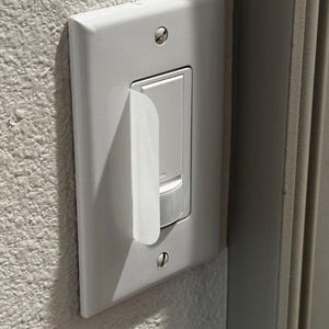 May include: A white rectangular light switch cover plate with a vertical rocker switch and a small motion sensor at the bottom. A thin, white, semi-transparent adhesive film strip is partially peeled away from the left side of the switch.