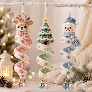 May include: Three hanging crochet ornaments featuring a reindeer with a pink scarf, a green Christmas tree with a star, and a snowman with a blue scarf. Each ornament has a spiraling base decorated with small beads and two metallic bells at the bottom.