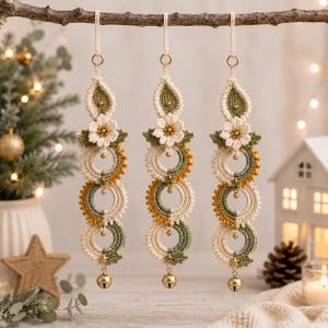 May include: Three hanging ornaments featuring cream, sage green, and mustard yellow crochet work. Each piece includes a central flower, three stacked circular rings, and a small gold-toned metal bell at the bottom, suspended by a matching cord.