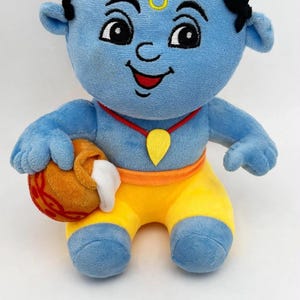 May include: A blue plush doll with black hair, wearing yellow pants and a red necklace with a yellow pendant. The doll holds a brown pot with white contents. A metal ruler is placed in front, showing the doll is approximately 9.8 inches tall.