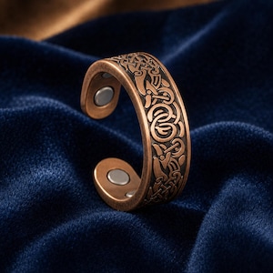 Könnte beinhalten: Ein kupferfarbener, offener Ring mit einem komplizierten, geprägten Knotenmuster auf der Außenseite. Die Innenseite des Rings zeigt zwei kleine, kreisförmige, silberfarbene magnetische Einsätze an beiden Enden des offenen Rings.