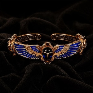 May include: A gold-toned cuff bracelet featuring a central dark blue scarab beetle motif flanked by outstretched wings with blue enamel detailing. The metalwork displays intricate engraved patterns and two small snake figures.